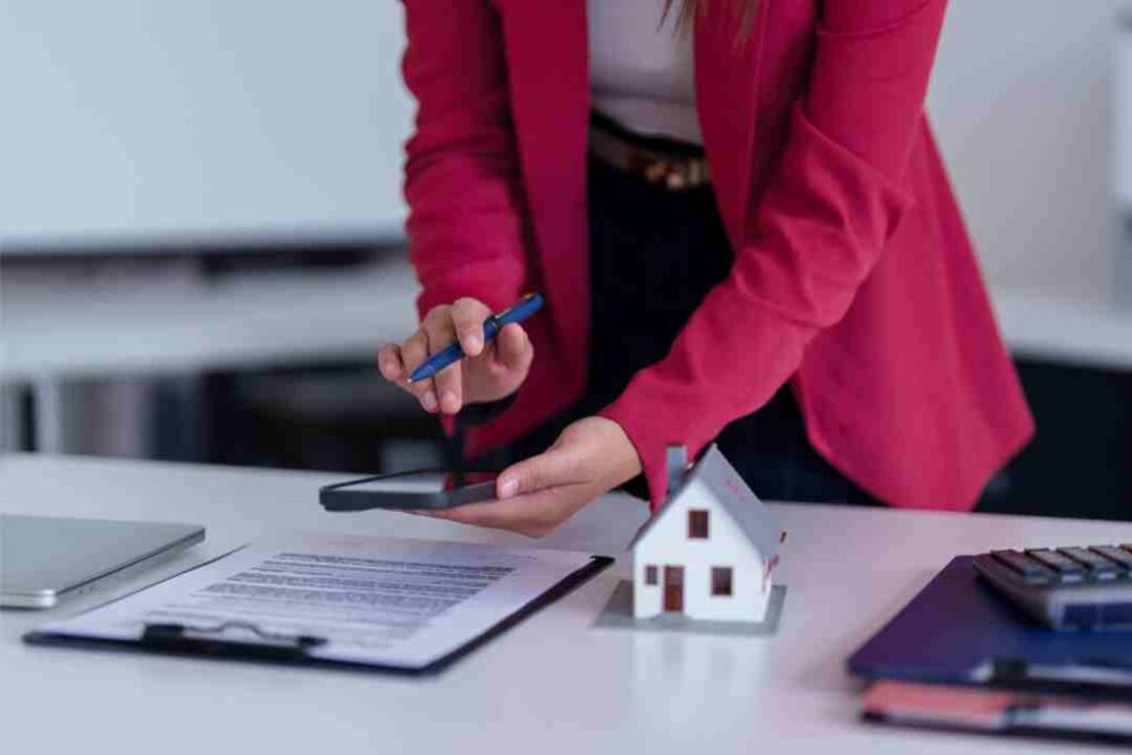 Person in pink blazer using a tablet with a contract, calculator, and house model, for real estate.