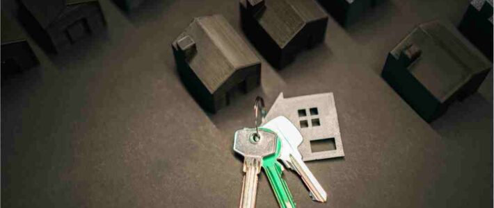 House keys and a house silhouette on a dark surface, with miniature houses in the background.